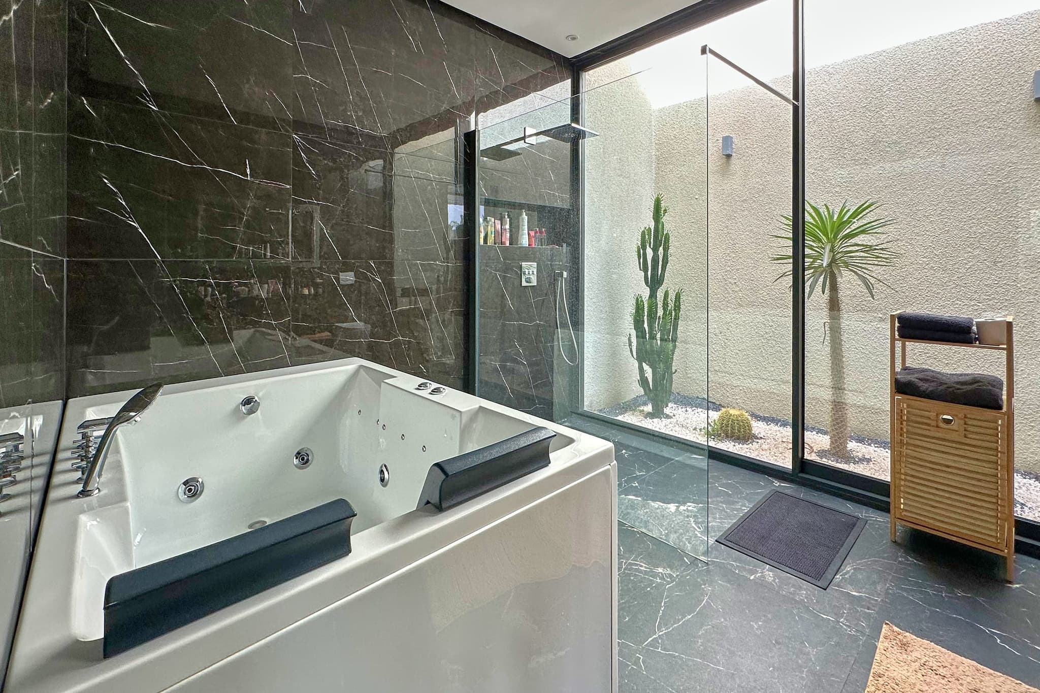 La salle de bain de la suite : marbre noir, baignoire balnéo, paroi vitrée sur un patio planté de cactus.