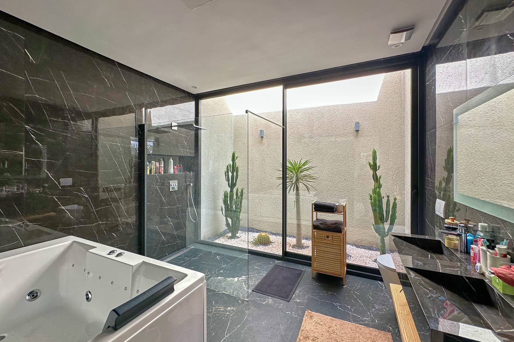 La même salle de bain vue en longueur : douche à l’italienne, baignoire, et le patio derrière la vitre.