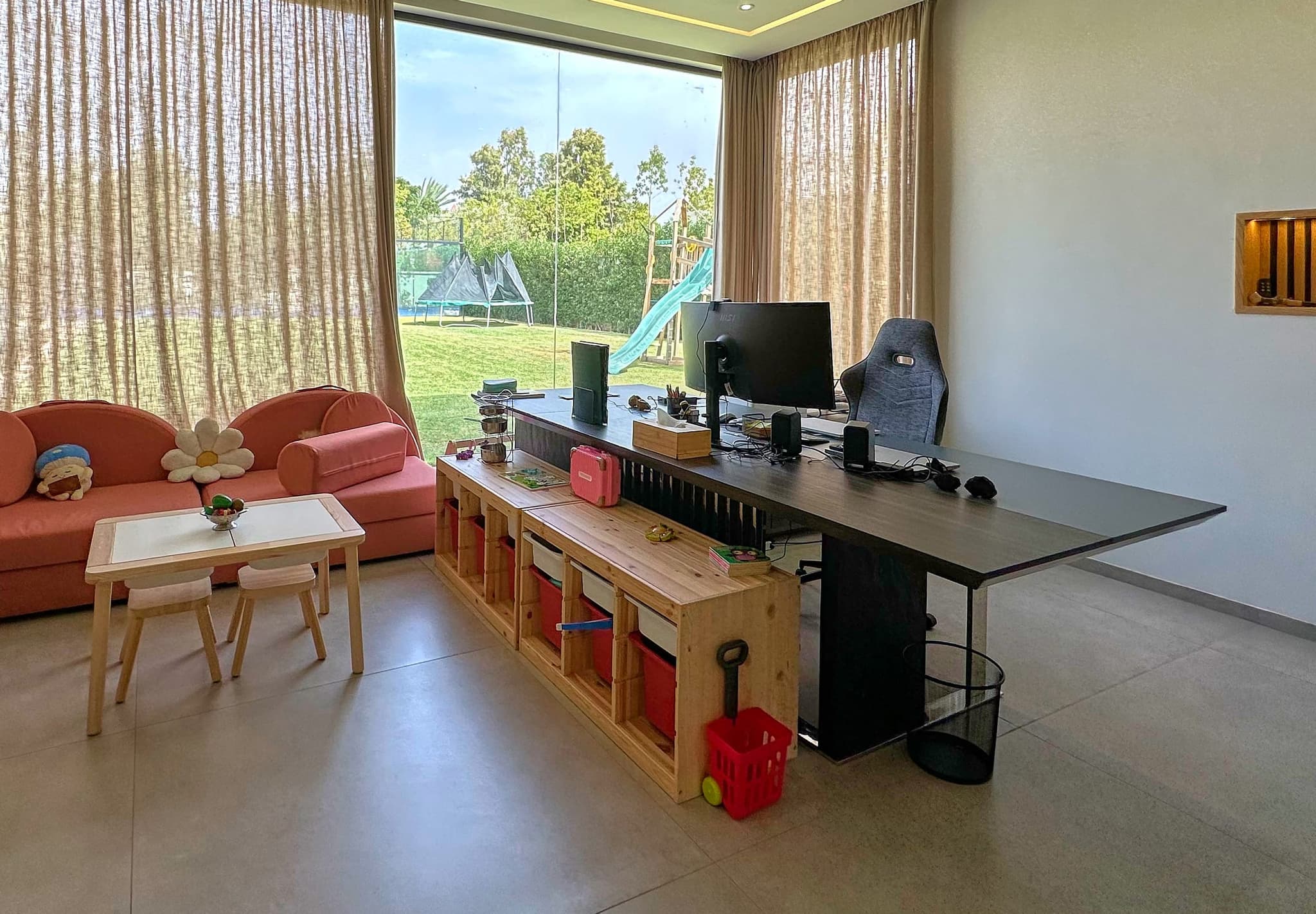 Le poste de travail : long plateau noir, écran et fauteuil, devant une baie vitrée ouverte sur le jardin et l’aire de jeux.