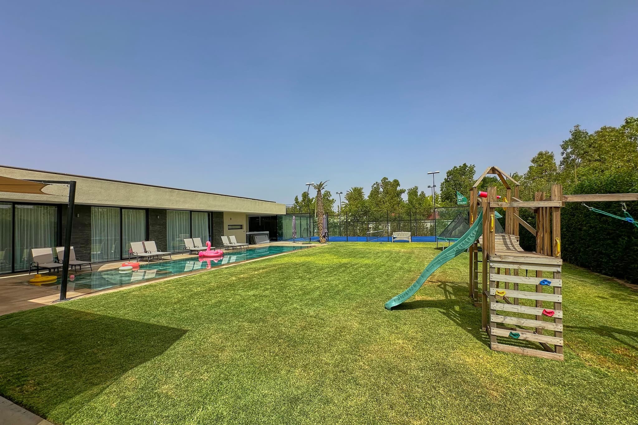 L’aire de jeux : portique en bois avec toboggan et trampoline, la maison et la piscine derrière.