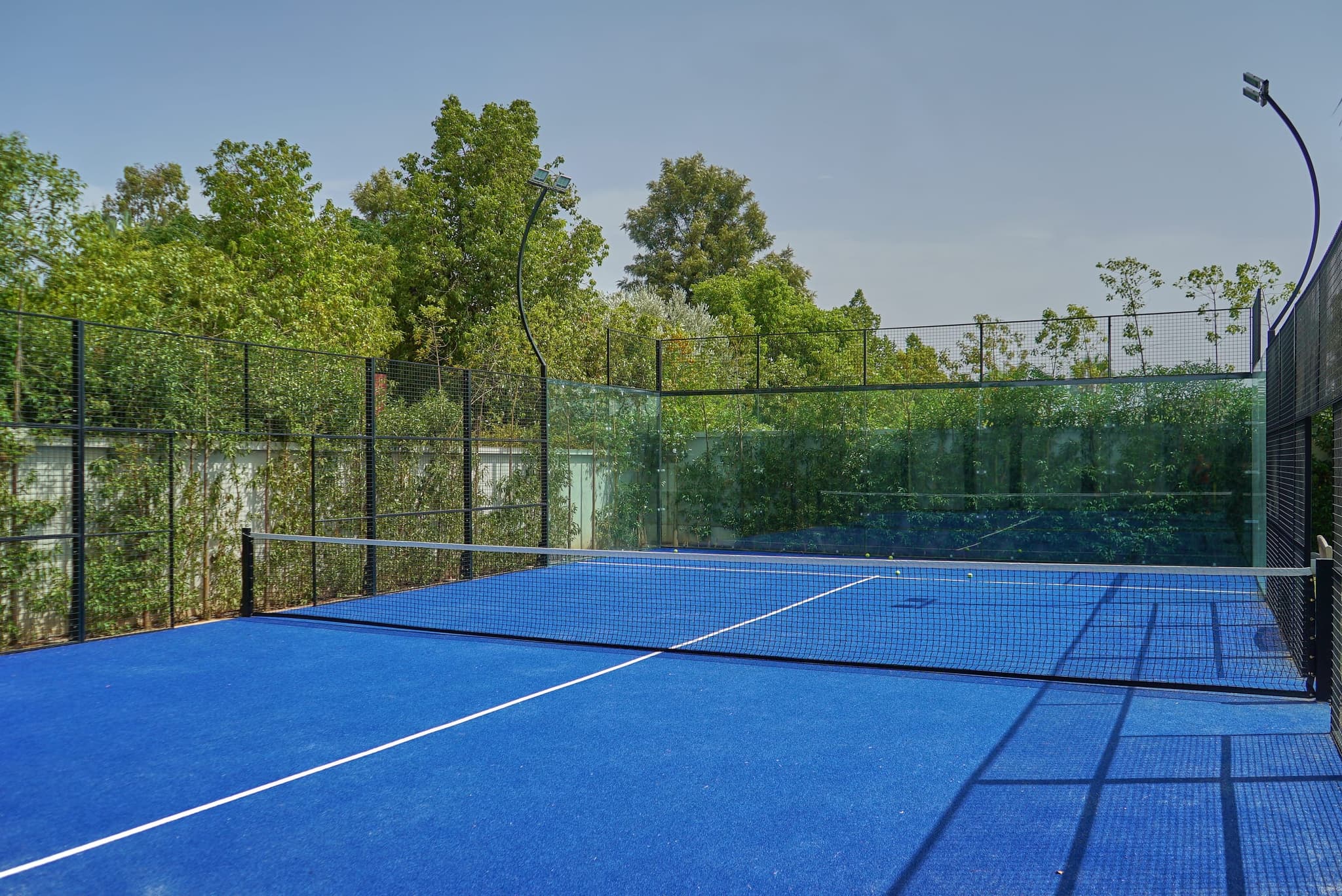 Le terrain de padel : surface bleue, filet, parois vitrées, et les deux projecteurs sur leurs mâts courbes.