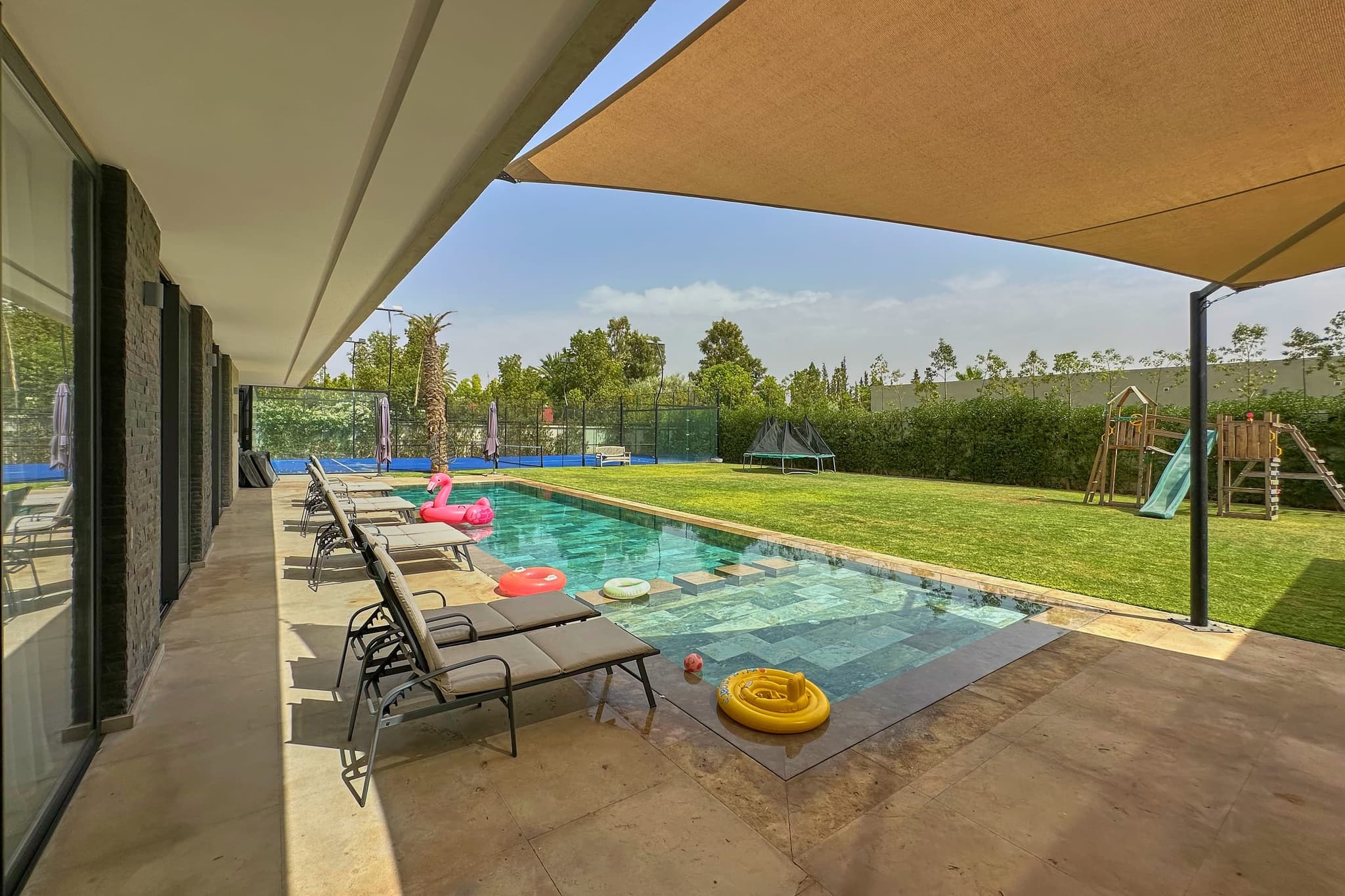 La piscine vue depuis la terrasse ombragée, transats alignés le long de la margelle, pelouse et aire de jeux au fond.