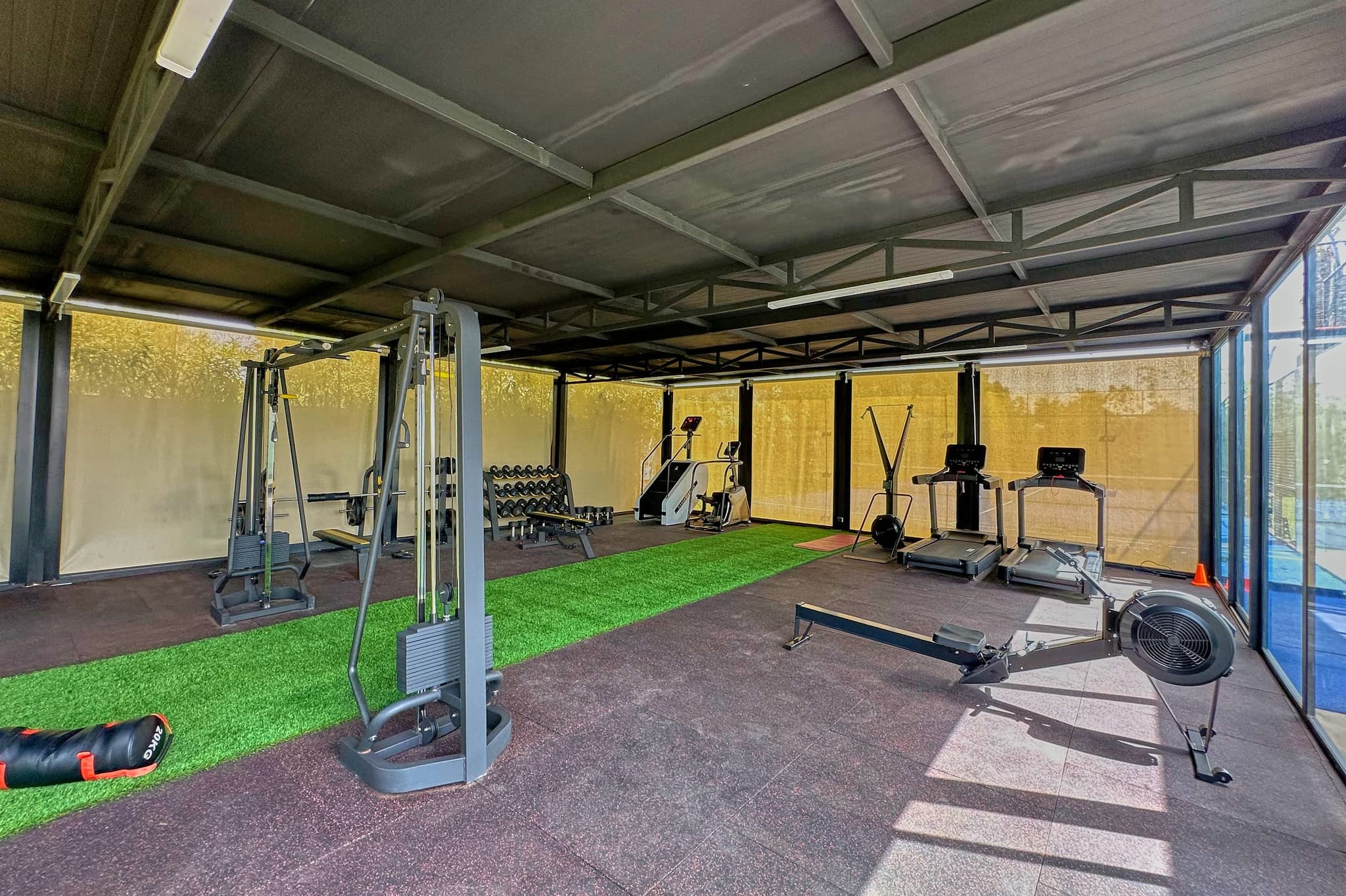 La salle de sport dans sa longueur : machines à charge guidée, tapis de course, rameur, gazon synthétique, vitrage sur le jardin.